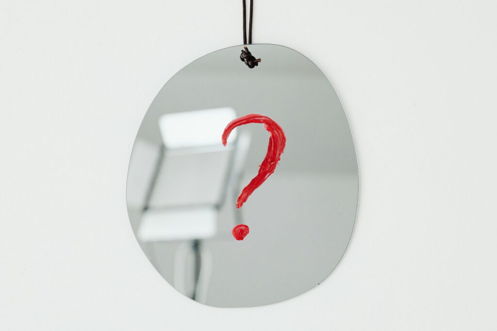 An oval mirror with a red question mark painted on it, hanging against a neutral background.